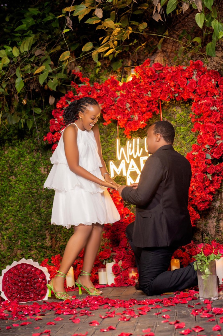 Brian on one knee holding Angella's hand in front of a heart of red roses and a neon “will you marry me” sign